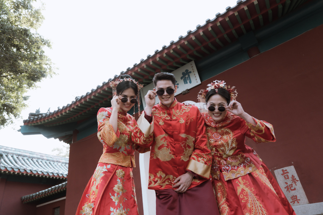 Three people in traditional red Chinese wedding attire, all wearing round sunglasses and grinning, in front of a red-and-gold temple rooftop.
