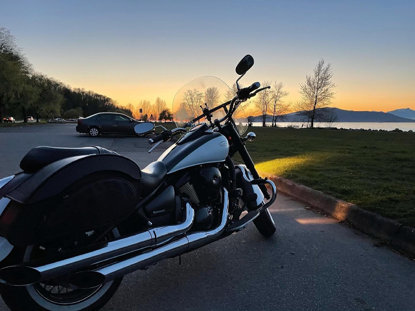 A Kawasaki cruiser motorcycle parked at a seaside lot at sunset, mountains and sky in the background.