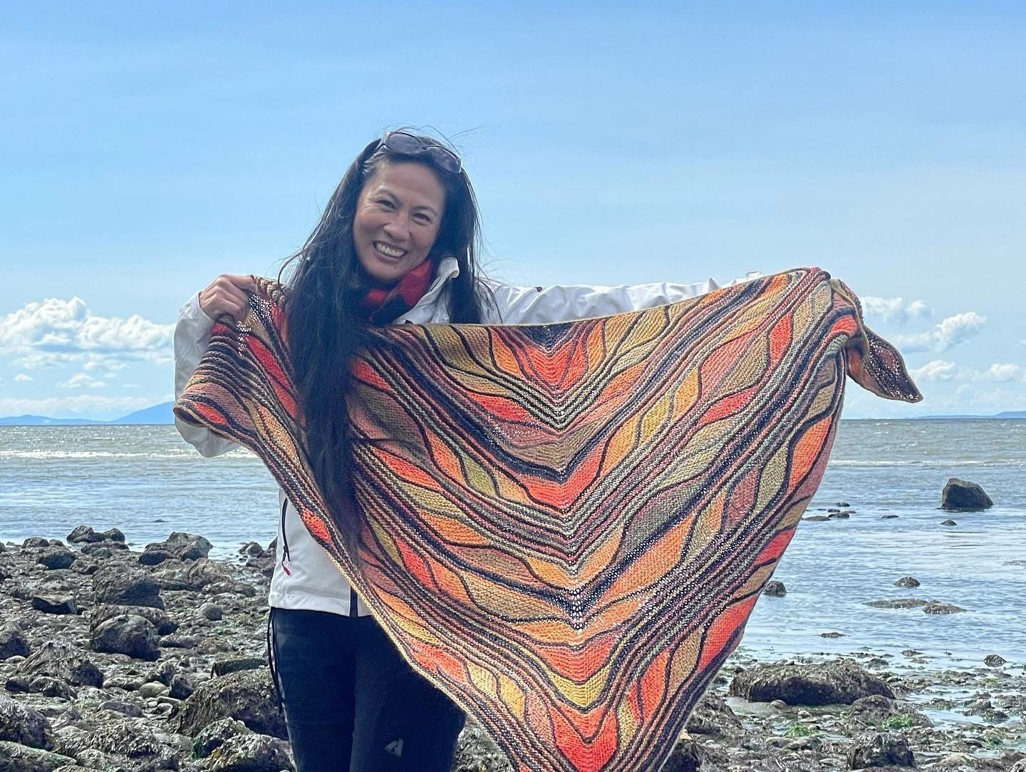 Gina on a rocky beach, smiling and holding up a large handknit triangular shawl in flame oranges, reds, and golds.